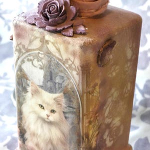 Puede incluir: Una botella rectangular con un acabado desgastado en tonos beige y marrón, que presenta una imagen de decoupage de un gato blanco de pelo largo dentro de un marco arqueado. La parte superior tiene una rosa de tela malva y tiras de tela clara.