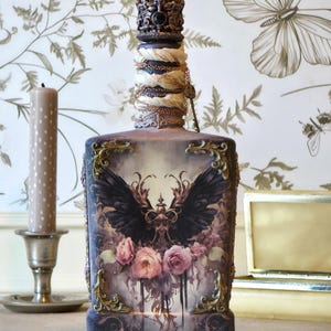 Może przedstawiać: Prostokątna szklana butelka z ciemną grafiką przedstawiającą czarne skrzydła i różowe róże. Butelka ozdobiona jest złotymi filigranowymi narożnikami, korkiem w kształcie korony oraz szyjką owiniętą kremowym materiałem i cienkimi złotymi łańcuszkami.