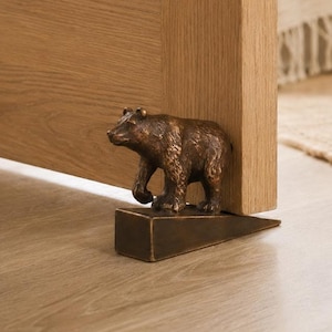 May include: A bronze-toned metal doorstop featuring a sculpted bear figure standing on a wedge-shaped base. The bear is depicted in a walking stance with textured fur details, positioned to hold a wooden door open on a light-coloured floor.