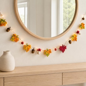 Puede incluir: Una guirnalda de luces decorativa con elementos de lana fieltrada que incluyen hojas de arce amarillas, naranjas y rojas, bellotas marrones, pequeñas bayas rojas y setas rojas con sombrero blanco, iluminada por pequeñas luces LED.