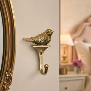 Puede incluir: Un gancho de pared de metal en tono dorado con un pájaro esculpido posado sobre una placa de montaje plana. El gancho tiene un diseño curvo con una punta redondeada y está asegurado a la pared con un tornillo visible. Acabado metálico.