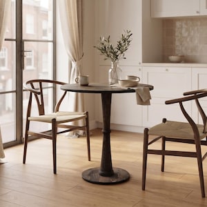 May include: A dark wood round pedestal table paired with two dark wood chairs featuring curved backrests and woven seats. The table is set with two ceramic mugs, a glass vase containing green leafy branches, and a small bowl with a linen napkin.