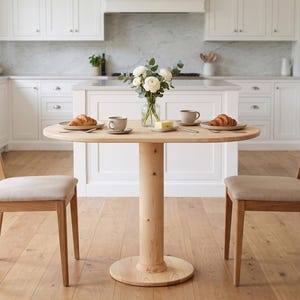 Op de afbeelding: Een ronde eettafel van lichtgekleurd hout met een centrale poot en twee bijpassende stoelen met beige gestoffeerde zittingen. De tafel is gedekt met twee keramische mokken, twee borden met croissants, een glazen vaas met witte bloemen en boter.