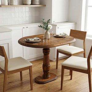 May include: An oval wooden dining table with a central pedestal base, accompanied by three chairs featuring wooden frames and off-white upholstered seats. The table is set with two plates, napkins, glasses, and a vase containing green foliage.