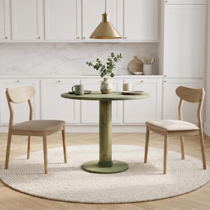 May include: A round green wooden pedestal dining table paired with two light wood chairs featuring beige upholstered seats. The table is set with a ceramic vase containing green foliage, two mugs, and two plates, all resting on a round woven rug.