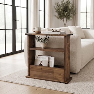 Puede incluir: Una mesa auxiliar rectangular de madera marrón oscuro con una superficie superior plana, un estante central y un compartimento de almacenamiento inferior. La mesa sostiene una taza de cerámica, un libro titulado KINIFOLK, una planta y dos revistas.