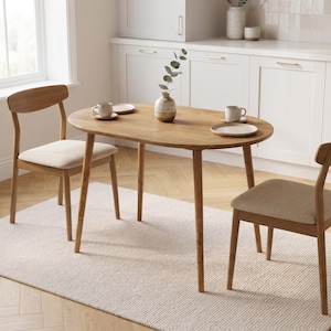 May include: An oval wooden dining table with four tapered legs and two matching wooden chairs featuring upholstered seats. The table is set with two ceramic plates, two mugs, and a small ceramic vase containing a green leafy stem.