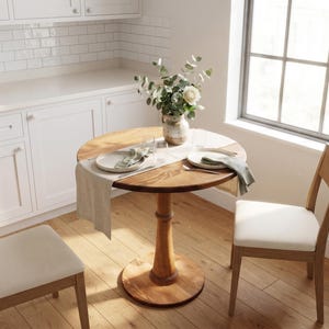 May include: A round wooden pedestal table with a light-colored fabric runner, two place settings with plates and napkins, and a ceramic vase containing greenery. Two wooden chairs with white upholstered seats are positioned at the table.