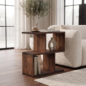 Puede incluir: Una mesa auxiliar de madera de color marrón oscuro con un perfil en forma de S que presenta dos compartimentos de almacenamiento abiertos. La superficie superior sostiene una taza de cerámica y un libro con gafas, mientras que el estante central contiene un pequeño jarrón blanco y la parte inferior contiene libros.
