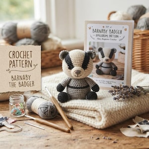 May include: A crochet pattern for Barnaby the Badger, featuring a small crocheted badger toy, a printed instruction booklet, two wooden crochet hooks, a ball of grey yarn, and a small glass jar with colourful stitch markers on a cream blanket.