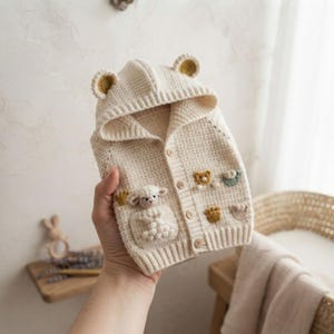 May include: A cream-colored, long-sleeved, hooded knit cardigan featuring a textured waffle stitch pattern. The garment includes a hood with small ears, a front pocket adorned with a crocheted sheep, and several small, colorful crocheted appliques.
