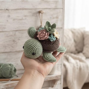 Pode incluir: Um brinquedo de tartaruga em crochê com uma carapaça castanho-escura adornada com várias pequenas suculentas de tecido macio em tons de rosa seco, verde sálvia e creme. A tartaruga tem uma cabeça verde-clara com olhos pretos e barbatanas.