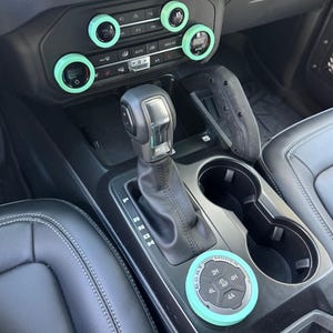 May include: A vehicle interior center console featuring a black gear shifter, two cup holders, and a circular G.O.A.T. modes dial. The climate control knobs and the drive mode dial are accented with bright mint green circular trim rings.
