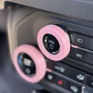 May include: Two circular light pink covers attached to the volume and temperature control knobs on a vehicle dashboard. The top knob features the text VOL and a power symbol, while the bottom knob displays a temperature reading in black and white.