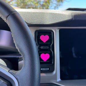 May include: Two bright pink heart-shaped air vent clips attached to the vertical slats of a vehicle's dashboard air conditioning vent. The clips are positioned one above the other, featuring a solid, matte finish and a simple, symmetrical design.