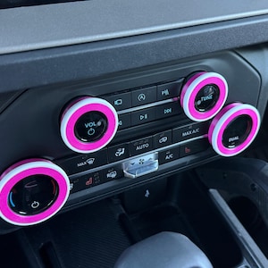 May include: A vehicle center console control panel featuring four circular knobs accented with bright pink rings. The black knobs include white text labels for VOL, TUNE, and DUAL functions, alongside various climate control buttons and icons.