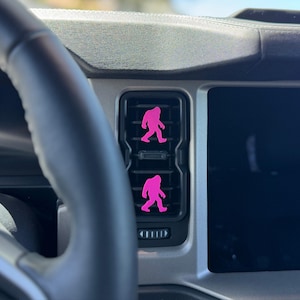 May include: Two bright pink Bigfoot silhouette decals are attached to the horizontal slats of a rectangular black car air vent. The vent is positioned on a silver dashboard next to a large black infotainment screen and a black steering wheel.