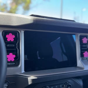 May include: Four bright pink flower-shaped air vent clips attached to the horizontal slats of a vehicle's dashboard air conditioning vents, positioned on either side of the central infotainment touchscreen display screen.