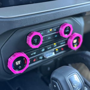 May include: Three bright pink, ridged circular covers installed over the climate control and volume knobs on a vehicle dashboard. The knobs feature digital displays showing temperature settings and volume icons, set against a dark gray console.