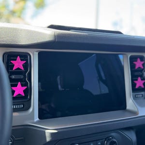 May include: Four bright pink star-shaped clips attached to the black air conditioning vents on the dashboard of a vehicle. The dashboard features a large central infotainment screen positioned between the two vertical vent panels.