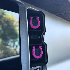 May include: Two small, bright pink horseshoe-shaped air vent clips with black textured centers are attached to the vertical slats of a dark gray vehicle air conditioning vent, positioned one above the other within the dashboard console.