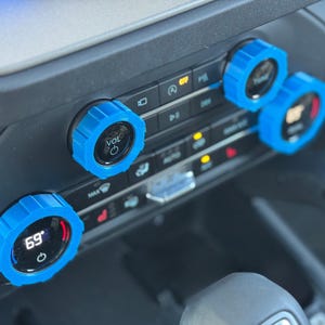 May include: Three bright blue, ridged circular covers installed over the climate control and volume knobs on a vehicle dashboard. The covers feature a textured, gear-like edge and fit over the existing black dials, which display digital settings.