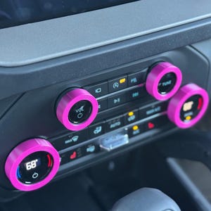 May include: A set of bright pink circular knob covers installed on the climate control and audio dials of a vehicle dashboard. The covers feature a smooth finish and surround the black dials, which display digital text including VOL, TUNE, and 68.