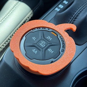 May include: An orange, pumpkin-shaped cover for a vehicle's G.O.A.T. Modes dial. The cover features a textured surface and fits over the black circular control knob, which displays settings for 2H, 4A, 4H, and 4L, along with a central icon.