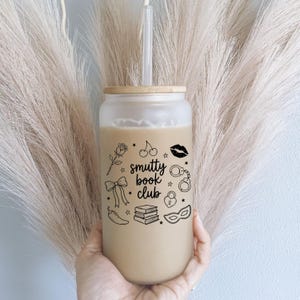 May include: A frosted glass tumbler with a bamboo lid and a clear straw, filled with a beige liquid. The exterior features black line art illustrations of a rose, cherries, lips, handcuffs, a bow, a padlock, a chili pepper, books, and a mask, with the text "smutty book club" in the center.