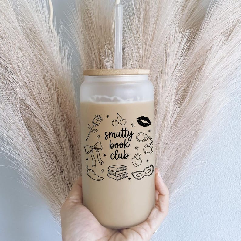 May include: A frosted glass tumbler with a bamboo lid and a clear straw, filled with a beige liquid. The exterior features black line art illustrations of a rose, cherries, lips, handcuffs, a bow, a padlock, a chili pepper, books, and a mask, with the text "smutty book club" in the center.