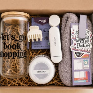 May include: A gift box containing a glass tumbler with a bamboo lid and the text 'let's go book shopping', a white hair claw, a box of Yogi tea, a white Vavolo device, a Capri Blue Volcano candle, a grey fuzzy sock, and a purple bookmark with a sticker.
