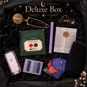 May include: A Deluxe Box gift set containing a gold tea infuser, a black velvet scrunchie, a Vahdam Assam Exotic tea pouch, a purple pen, a white envelope with a gold wax seal, a white portable light, sticky notes, and decorative stickers.
