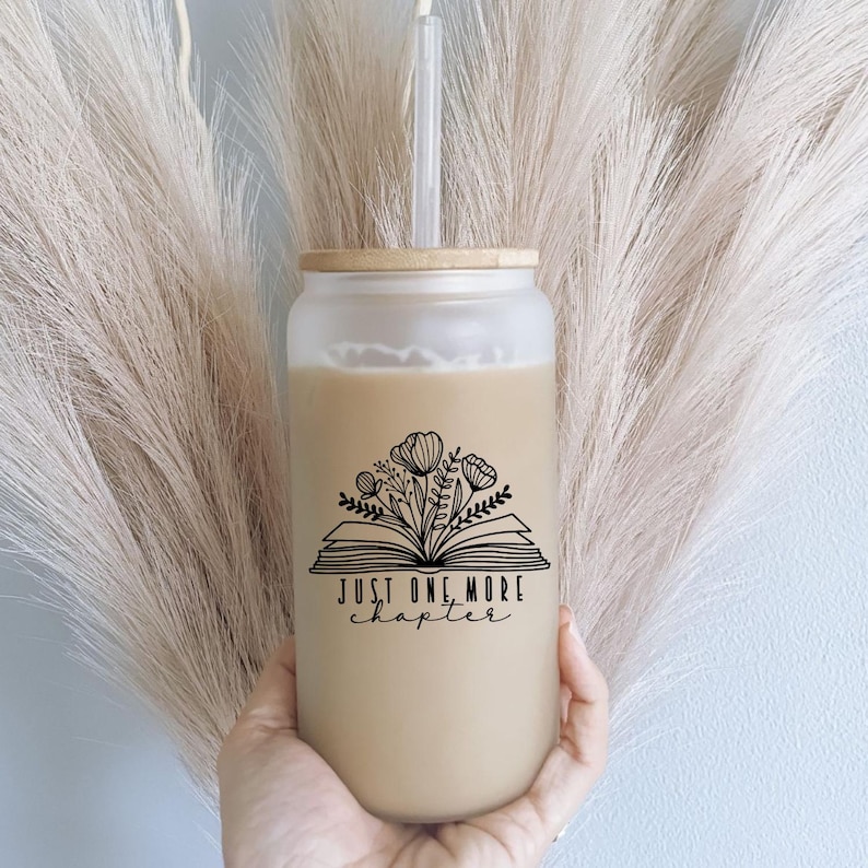 May include: A frosted glass tumbler with a bamboo lid and a clear straw, filled with a light brown beverage. The glass features a black line art graphic of an open book with flowers emerging from the pages and the text Just One More Chapter.