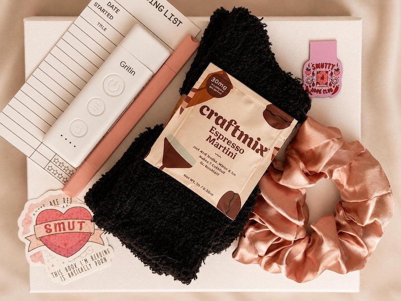 May include: A gift set containing a white reading list notepad, a white Gritin book light, a pink pen, black fuzzy socks, a Craftmix Espresso Martini drink packet, a pink satin scrunchie, and two stickers with text including Smutty Book Club.