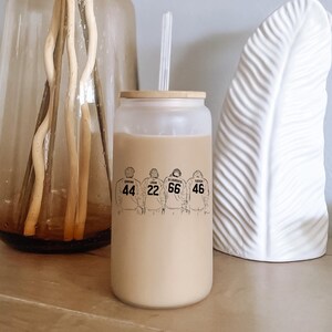 May include: A frosted glass tumbler filled with a light brown beverage, featuring a bamboo lid and a clear straw. The exterior displays a black line art graphic of four hockey players wearing jerseys with the names Graham, Logan, Di Laurentis, and Tucker.