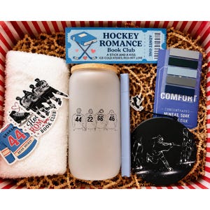 May include: A gift box containing a white towel with hockey player graphics, a frosted glass tumbler with four hockey jerseys numbered 44, 22, 66, and 46, a light blue pen, a Hockey Romance Book Club ticket, a Comfort soak packet, and a black hockey coaster.