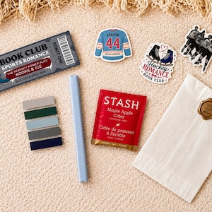 May include: A collection of items including a rectangular ticket reading Book Club Sports Romance, three stickers featuring a hockey jersey, skates, and players, a light blue marker, a set of five colored page flags, a Stash tea bag, and a white envelope.