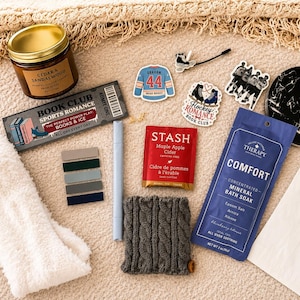 May include: A gift set including a Cedar & Sandalwood candle, a Book Club Sports Romance bookmark, a blue hockey jersey sticker, a Hockey Romance sticker, a Stash Maple Apple Cider tea bag, Comfort mineral bath soak, and white fuzzy socks.