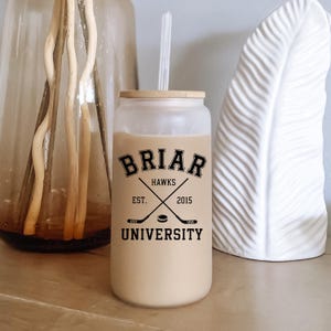 May include: A frosted glass tumbler with a bamboo lid and a clear straw, filled with a light brown beverage. The exterior features black text reading BRIAR UNIVERSITY, HAWKS, EST. 2015, and a graphic of two crossed hockey sticks and a puck.