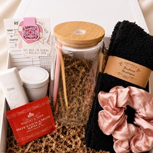 May include: A gift box containing a glass tumbler with a bamboo lid, a pink pen, a white Gritin device, a Harney & Sons Hot Cinnamon Spice tea packet, a black fuzzy pair of socks with a Fashion Boutique label, and a pink satin scrunchie.