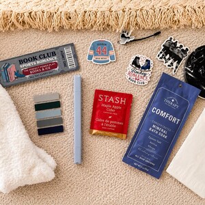 May include: A collection of items including a pair of white fuzzy socks, a grey marker, a Stash Maple Apple Cider tea bag, a blue Comfort mineral bath soak packet, a book club bookmark, hockey-themed stickers, and a black circular coaster.