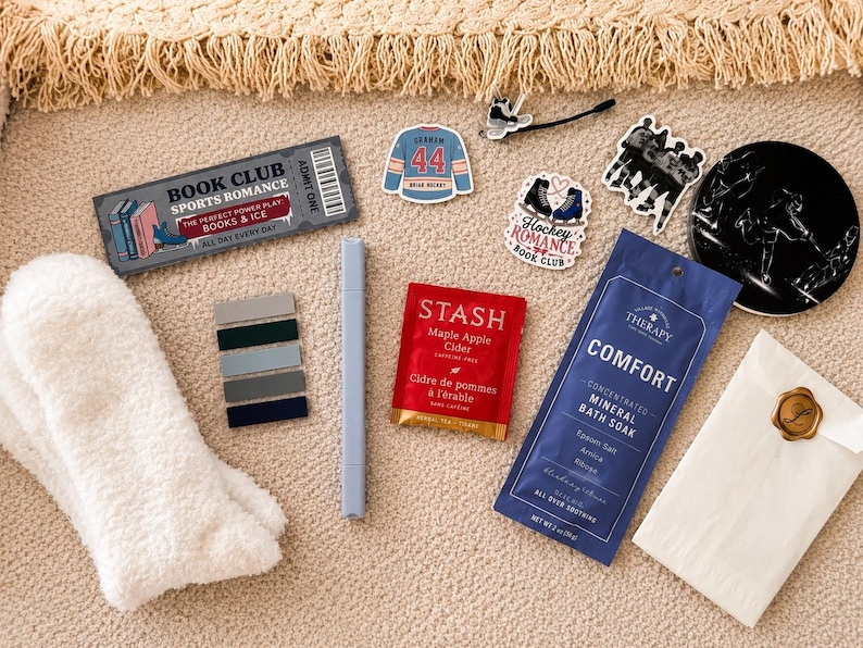 May include: A collection of items including a pair of white fuzzy socks, a grey marker, a Stash Maple Apple Cider tea bag, a blue Comfort mineral bath soak packet, a book club bookmark, hockey-themed stickers, and a black circular coaster.