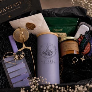 May include: A gift box containing a lavender canister labeled Velaris City of Starlight, a gold tea infuser, a candle, a green tea packet, a white envelope with a gold wax seal, a purple pen, a set of purple sticky notes, and a black scrunchie.