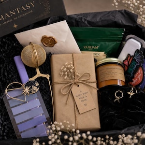 May include: A gift box containing a book wrapped in brown paper with a tag reading A BLIND DATE WITH A BOOK, a gold tea infuser, a scented candle, a purple bookmark, a white envelope with a gold wax seal, a green tea pouch, and black accessories.