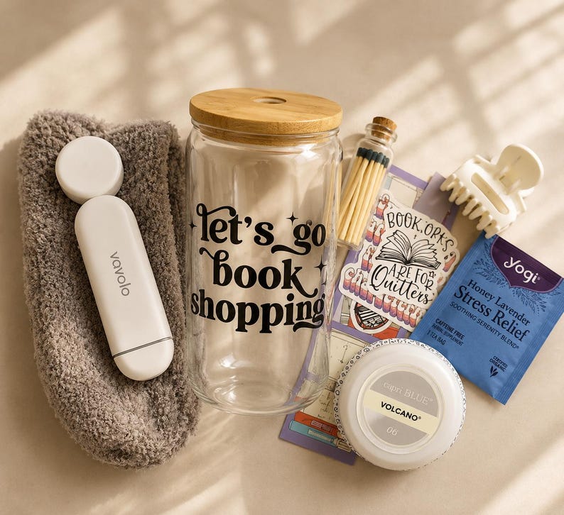 May include: A gift set containing a fuzzy brown sock, a white Vavolo device, a clear glass tumbler with a bamboo lid and the black text "let's go book shopping", a small glass vial of matches, a sticker, a white hair claw, and a Yogi tea bag.