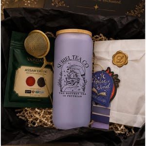 May include: A frosted lavender glass tumbler with a bamboo lid, featuring black text reading Suriel Tea Co and The Hottest Tea in Prythian. Includes a gold metal tea infuser, a green tea pouch, and a white paper envelope with a gold wax seal.