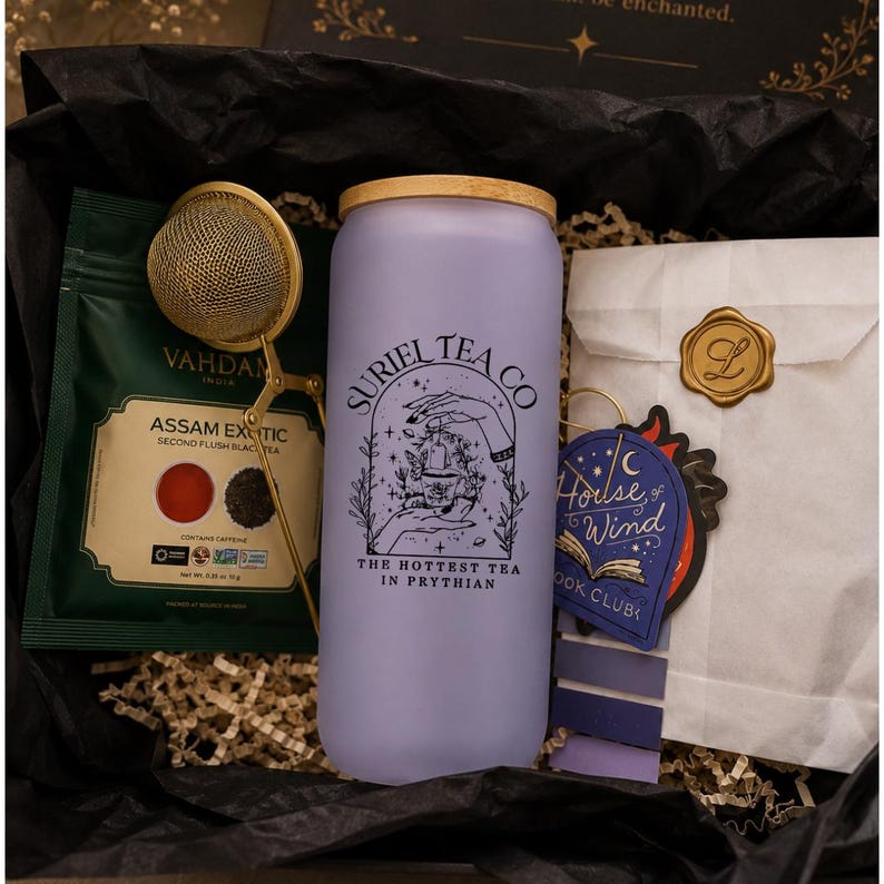 May include: A frosted lavender glass tumbler with a bamboo lid, featuring black text reading Suriel Tea Co and The Hottest Tea in Prythian. Includes a gold metal tea infuser, a green tea pouch, and a white paper envelope with a gold wax seal.