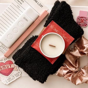 May include: A white reading light with the brand name Gritin, a pink pen, a black fuzzy sock, a candle in a white container with a red label, a pink satin scrunchie, a notepad, and two stickers, one shaped like a heart with the text SMUT.
