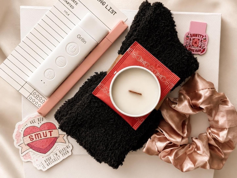 May include: A white reading light with the brand name Gritin, a pink pen, a black fuzzy sock, a candle in a white container with a red label, a pink satin scrunchie, a notepad, and two stickers, one shaped like a heart with the text SMUT.