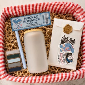 May include: A gift box containing a frosted glass tumbler with a bamboo lid, a light blue pen, a rectangular card labeled Hockey Romance Book Club, a set of five adhesive page markers, and a white envelope sealed with a gold wax L stamp.